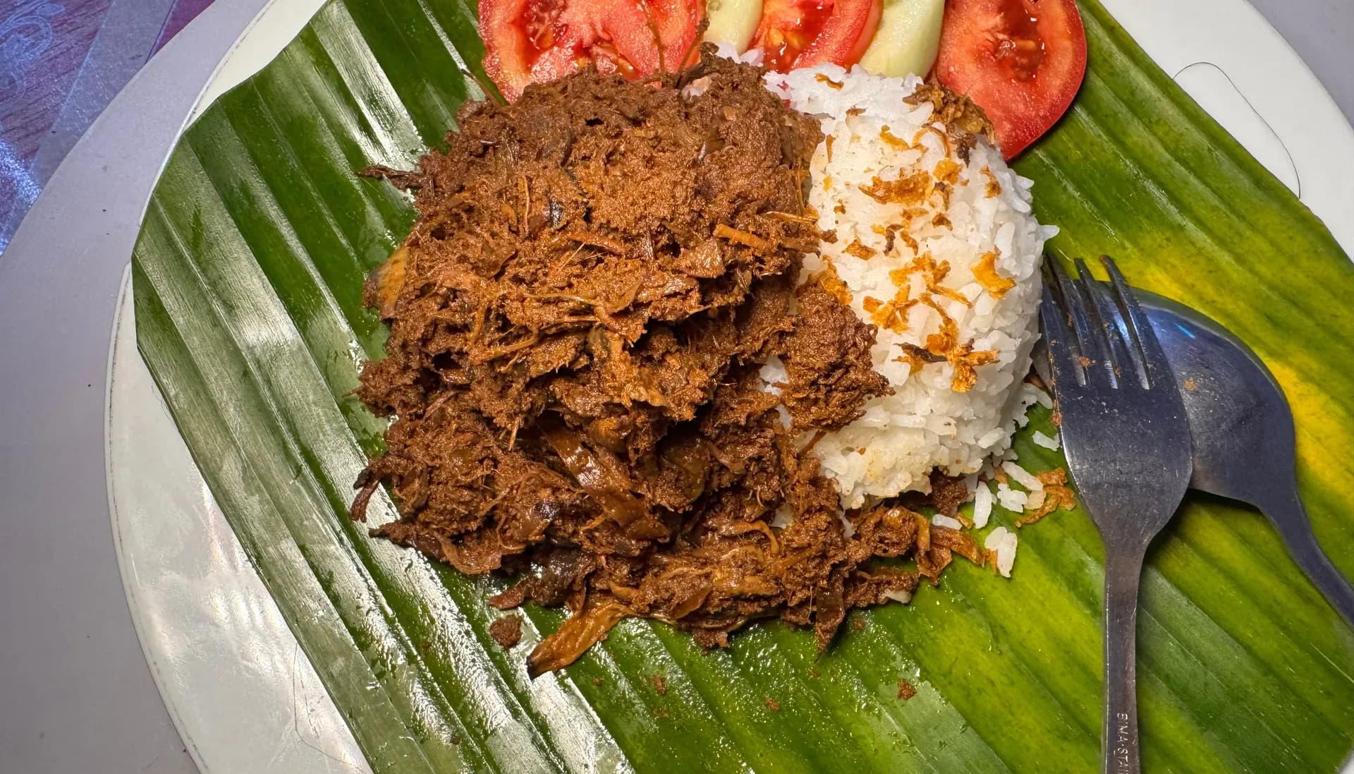 Rendang jantung pisang dengan nasi putih, tomat, dan timun di atas daun pisang