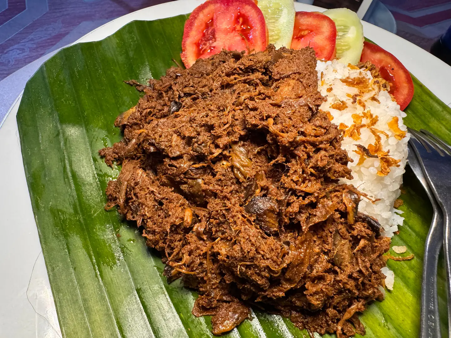 Rendang jantung pisang dari dekat di atas daun pisang, dengan nasi dan irisan tomat
